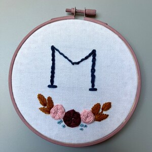 Könnte beinhalten: Stickerei-Kunst auf einem Reifen mit dem Buchstaben "M" in marineblauem Garn, umgeben von einem Kranz aus braunen Blättern und rosa und burgunderfarbenen Blumen.