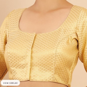 Könnte beinhalten: Eine goldene Brokatbluse mit rundem Ausschnitt und Knopfleiste. Die Bluse hat kurze Ärmel und ein geometrisches Muster.