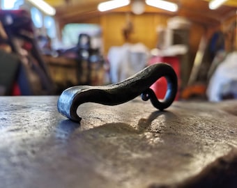 Può includere: Un apribottiglie in ferro battuto a mano con una finitura scura e testurizzata. L'oggetto presenta un corpo curvo e allungato che si assottiglia in una punta a un'estremità e termina con un ricciolo circolare stretto all'altra, metallo grezzo.