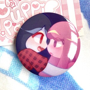May include: A round button featuring a cartoon illustration of two characters, one with dark blue hair and a red and black checked shirt, and the other with pink hair. The characters are embracing each other.