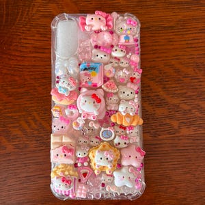 May include: A clear phone case adorned with numerous pink and white Hello Kitty-themed charms. The case features various designs, including Hello Kitty figures, bows, desserts, and other decorative elements. The overall aesthetic is cute and playful.