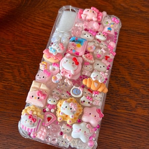 May include: Clear phone case decorated with pink Hello Kitty figures, bows, and food-themed charms. The case features a variety of 3D embellishments, including a clock, cake, and popcorn, creating a playful and decorative accessory.