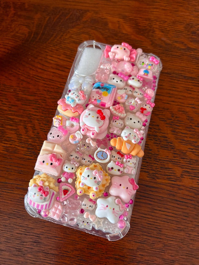 May include: Clear phone case decorated with pink Hello Kitty figures, bows, and food-themed charms. The case features a variety of 3D embellishments, including a clock, cake, and popcorn, creating a playful and decorative accessory.