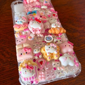 May include: A clear phone case adorned with numerous pink and white Hello Kitty-themed charms. The case features various 3D embellishments, including Hello Kitty figures, desserts, and other decorative elements. The overall design is playful and colorful.