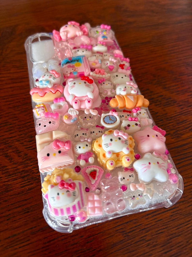 May include: A clear phone case adorned with numerous pink and white Hello Kitty-themed charms. The case features various 3D embellishments, including Hello Kitty figures, desserts, and other decorative elements. The overall design is playful and colorful.