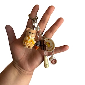 May include: A keychain featuring a miniature flocked bear figurine in a yellow outfit, a small chocolate bar charm, a tiny ice cream cone, a miniature plate with the word pudding, a small milk bottle, and a tiny coffee mug with latte art.
