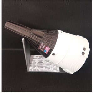 May include: A 3D printed model of the Gemini spacecraft, a white capsule with a brown and black base. The model is mounted on a stand and includes the text "UNITED STATES" and "GEMINI Lanceur Diamètre: 3.06m - Hauteur" along with a list of Gemini missions and crew members.