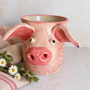May include: A handmade ceramic mug shaped like a pig, featuring a glossy light pink glaze, a prominent snout, and two large protruding ears. The mug has a smooth interior and a textured exterior surface with small, subtle sculpted details.