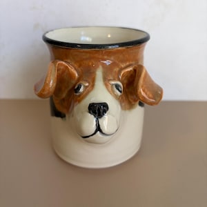 May include: A ceramic mug featuring a three-dimensional sculpted dog head with floppy ears. The dog has a white muzzle, brown patches around the eyes, and a black nose. The mug has a smooth, glossy finish with a dark rim and a cylindrical body shape.