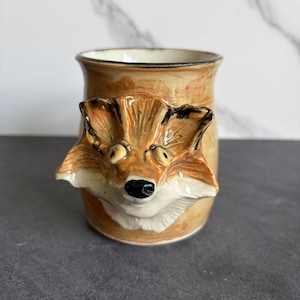 May include: A ceramic mug with a fox face design. The mug is tan with a detailed fox head in shades of orange, black, and white. The fox's face is sculpted and protrudes from the mug's surface, adding a 3D effect. The interior of the mug is a cream color.