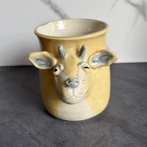 May include: A ceramic mug with a goat face design. The mug is primarily yellow with a cream-colored interior. The goat's face is detailed with gray accents on the ears, horns, and facial features. The mug has a slightly flared rim.