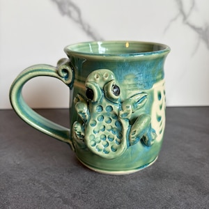 May include: A handmade ceramic mug with a teal glaze and a sculpted frog design. The mug features a curved handle and a cream-colored interior. The frog has black eyes and is textured with small circles.