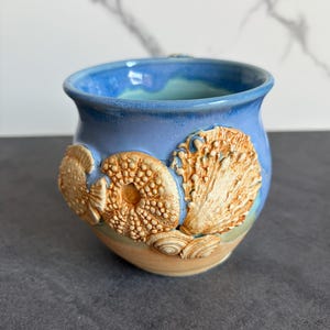 May include: A ceramic mug with a blue interior and exterior, adorned with raised seashell designs in shades of tan and cream. The mug has a beach-themed design, perfect for coastal decor.