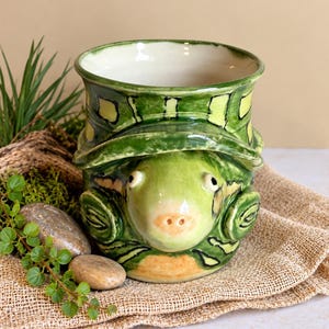 May include: A ceramic mug shaped like a turtle with a glossy green and yellow glaze. The mug features a sculpted turtle head protruding from the front, with dark outlines defining the shell pattern and a smooth, cream-colored interior.