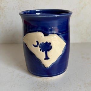 May include: A deep blue ceramic mug featuring a raised, cream-colored applique in the shape of the state of South Carolina. The applique displays a dark blue crescent moon and palmetto tree emblem centered on the state's geographic outline.