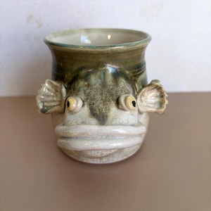 May include: A ceramic mug shaped like a fish head with a wide mouth, protruding eyes, and side fins. The mug features a mottled green and beige glaze with a smooth, glossy finish, showcasing a detailed sculpted face on the front exterior.