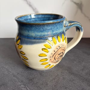 May include: Handmade ceramic mug with a blue rim and handle. The mug features a cream-colored body with a sunflower design in yellow and brown. The mug is a unique piece of pottery.
