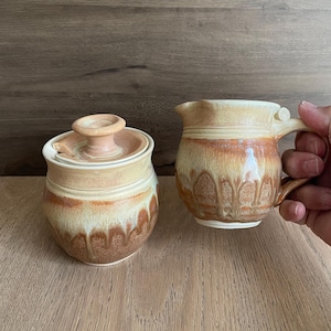 May include: A set of two ceramic sugar and creamer containers with a brown and white glaze. The containers have a unique, textured design.