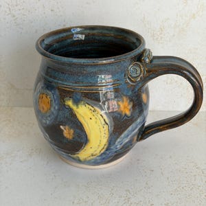 May include: A ceramic mug with a glossy dark blue and brown glaze featuring a hand-painted yellow crescent moon and small orange stars. The mug has a rounded body, a wide rim, and a curved handle with a small decorative spiral detail at the top.