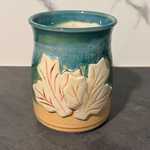 May include: A ceramic mug featuring a two-tone glaze with a deep teal blue top and a light tan base. The exterior is decorated with two raised, white, embossed maple leaf designs that have subtle red and orange accents on the leaf veins.