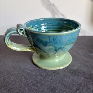 May include: A ceramic coffee pour-over dripper featuring a two-tone glaze with a deep blue upper section that transitions into a light sage green base. The piece includes a wide circular foot, a flared rim, and a single handle with a scroll detail.