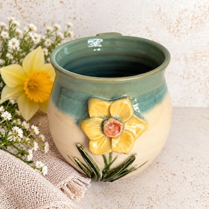 May include: A ceramic mug with a rounded body featuring a two-tone glaze of sage green on the upper portion and cream on the lower portion. A three-dimensional yellow daffodil with green leaves is attached to the front of the mug.