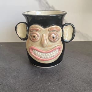 Puede incluir: Una taza de cerámica negra con un diseño de cara de mono. La cara del mono es de color canela con una amplia sonrisa de labios rosados y grandes dientes blancos. La taza tiene dos asas en forma de oreja de color canela y un interior color crema.