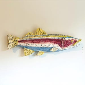 May include: A ceramic fish wall sculpture featuring a vibrant rainbow trout design. The body displays a prominent magenta lateral stripe, blue underbelly, and yellow fins, all covered in small black speckles. The fish has a detailed head.