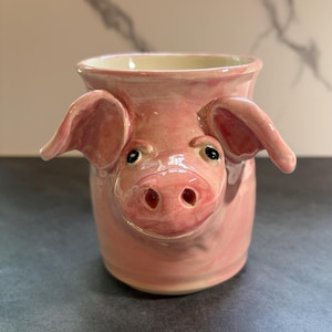 May include: A pink ceramic mug shaped like a pig's head. The mug features a three-dimensional snout and two protruding ears on the sides. The surface has a glossy finish with subtle variations in pink tones, and the interior is a smooth cream color.