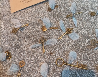 May include: A collection of decorative keys with delicate, translucent wings. The keys are made of gold-toned metal and have a vintage aesthetic. The wings are a light blue color and are made of a sheer fabric. The keys are arranged on a gray, textured surface. The text "Love" is visible on one of the keys.