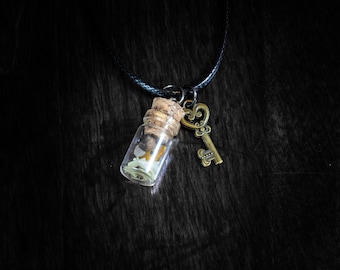 May include: A black cord necklace with a small glass bottle filled with white and brown objects. The bottle has a cork stopper and is attached to a gold-toned key charm.