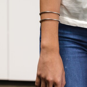 May include: Two silver-colored, thin, round bangles are worn on the forearm. The person is wearing a white top and blue jeans. The hand is relaxed, with the fingers slightly curved. The background is a white wall.