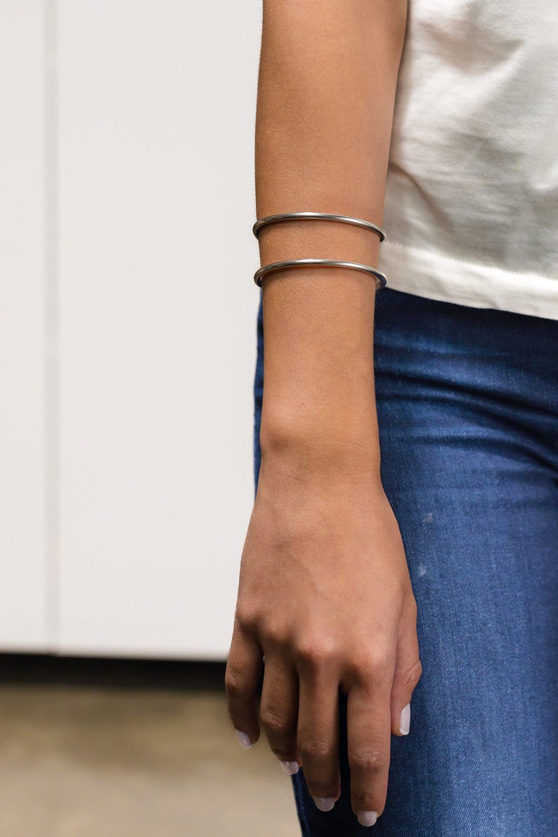 May include: Two silver-colored, thin, round bangles are worn on the forearm. The person is wearing a white top and blue jeans. The hand is relaxed, with the fingers slightly curved. The background is a white wall.