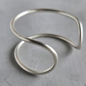 May include: A silver-colored cuff bracelet with a unique, flowing design. The bracelet curves in a continuous loop, creating an open, minimalist aesthetic. The metal has a polished finish, reflecting light. The overall style is modern and elegant.