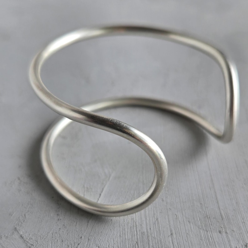 May include: A silver-colored cuff bracelet with a unique, flowing design. The bracelet curves in a continuous loop, creating an open, minimalist aesthetic. The metal has a polished finish, reflecting light. The overall style is modern and elegant.
