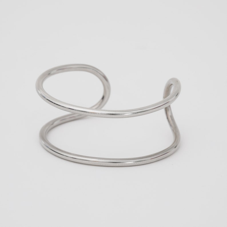May include: A silver-colored cuff bracelet with a minimalist design. The bracelet features a continuous, curved metal band that forms two parallel lines connected by a loop. The overall shape is open and modern.