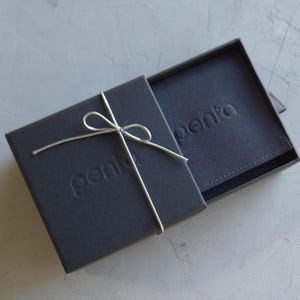 May include: A dark gray gift box with a matching interior, tied with a silver string bow. The word "penta" is embossed on both the box and the interior. The box is partially open, revealing the contents.