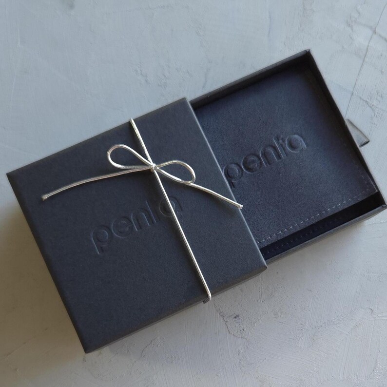 May include: A dark gray gift box with a matching interior, tied with a silver string bow. The word "penta" is embossed on both the box and the interior. The box is partially open, revealing the contents.