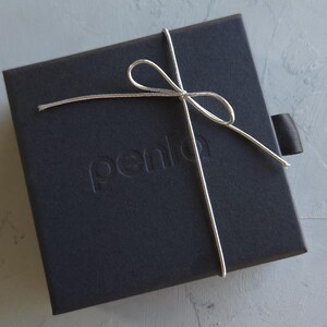 May include: A black square gift box tied with a silver string and bow. The box has the word "penlo" embossed on the top. A black ribbon is attached to the side of the box.