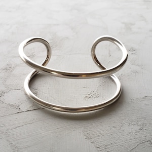 May include: A silver-colored cuff bracelet with a minimalist design. The bracelet features a lower, circular band and an upper band that curves into loops on each side. The jewelry is displayed on a textured, light gray surface.