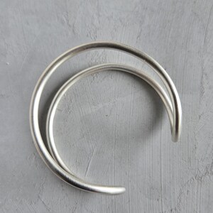 May include: A silver-colored, open cuff bracelet with a curved, minimalist design. The bracelet features a smooth, rounded band that forms a partial circle, creating a modern and elegant aesthetic. The open design allows for easy wear and adjustment.