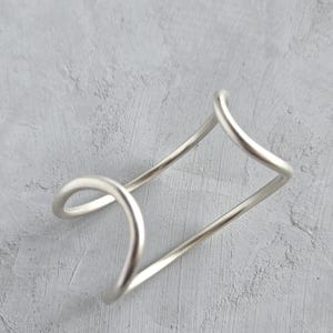 May include: A silver-colored cuff bracelet with a unique, open design. The bracelet features a geometric shape with curved and straight lines, creating an abstract and modern aesthetic. The metal has a matte finish, and the bracelet appears to be lightweight.
