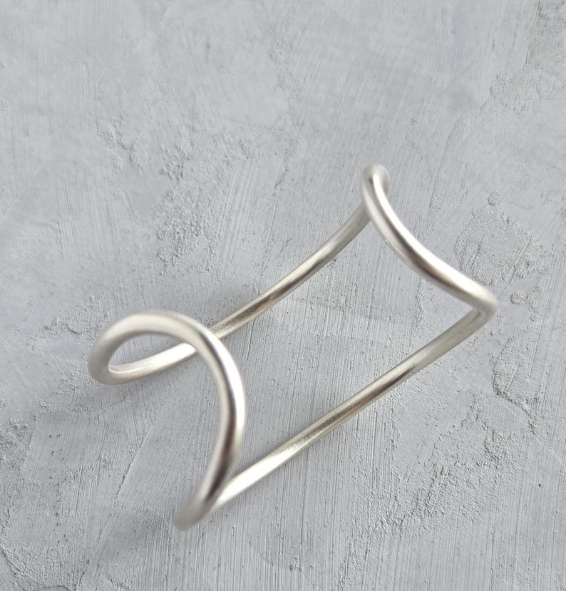 May include: A silver-colored cuff bracelet with a unique, open design. The bracelet features a geometric shape with curved and straight lines, creating an abstract and modern aesthetic. The metal has a matte finish, and the bracelet appears to be lightweight.