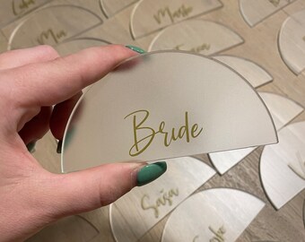 May include: Clear acrylic semi-circle place cards with gold lettering. One card is held up, displaying the word "Bride". Other cards with names are scattered on a wooden surface. Ideal for weddings and events.
