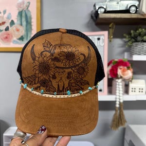 May include: A brown trucker hat with a black mesh back. The front panel has a laser-etched design of a bull skull, sunflowers, and leaves. A band of turquoise beads and gold studs decorates the brim. The hat has a curved brim and a button on top.
