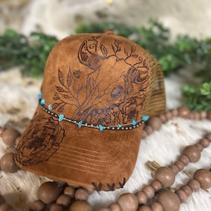 Puede incluir: Una gorra de camionero de gamuza marrón y malla con un diseño floral y de calavera. La gorra tiene un ribete de cuentas turquesa.