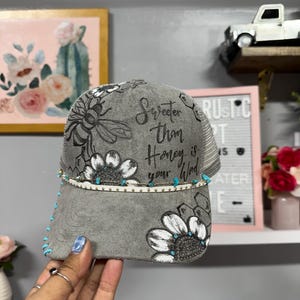 May include: Gray trucker hat with floral designs and the text "Sweeter Than Honey is your Word." The hat features a mesh back, turquoise accents, and a decorative band with small gold and turquoise beads.