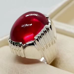 May include: A silver ring featuring a large, oval-shaped, deep red gemstone. The ring has a textured, ridged design on the sides, and the gemstone is polished to a smooth, glossy finish. The ring is set against a neutral background.