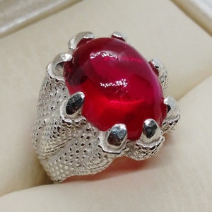 May include: A silver ring featuring a large, oval, red gemstone. The ring's design resembles a claw, with textured metal forming the band and gripping the stone. The gemstone has a glossy, polished appearance.