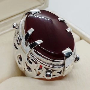 Könnte beinhalten: Ein silberner Ring mit einem verzierten Design und einem großen, ovalen, dunkelroten Edelstein.